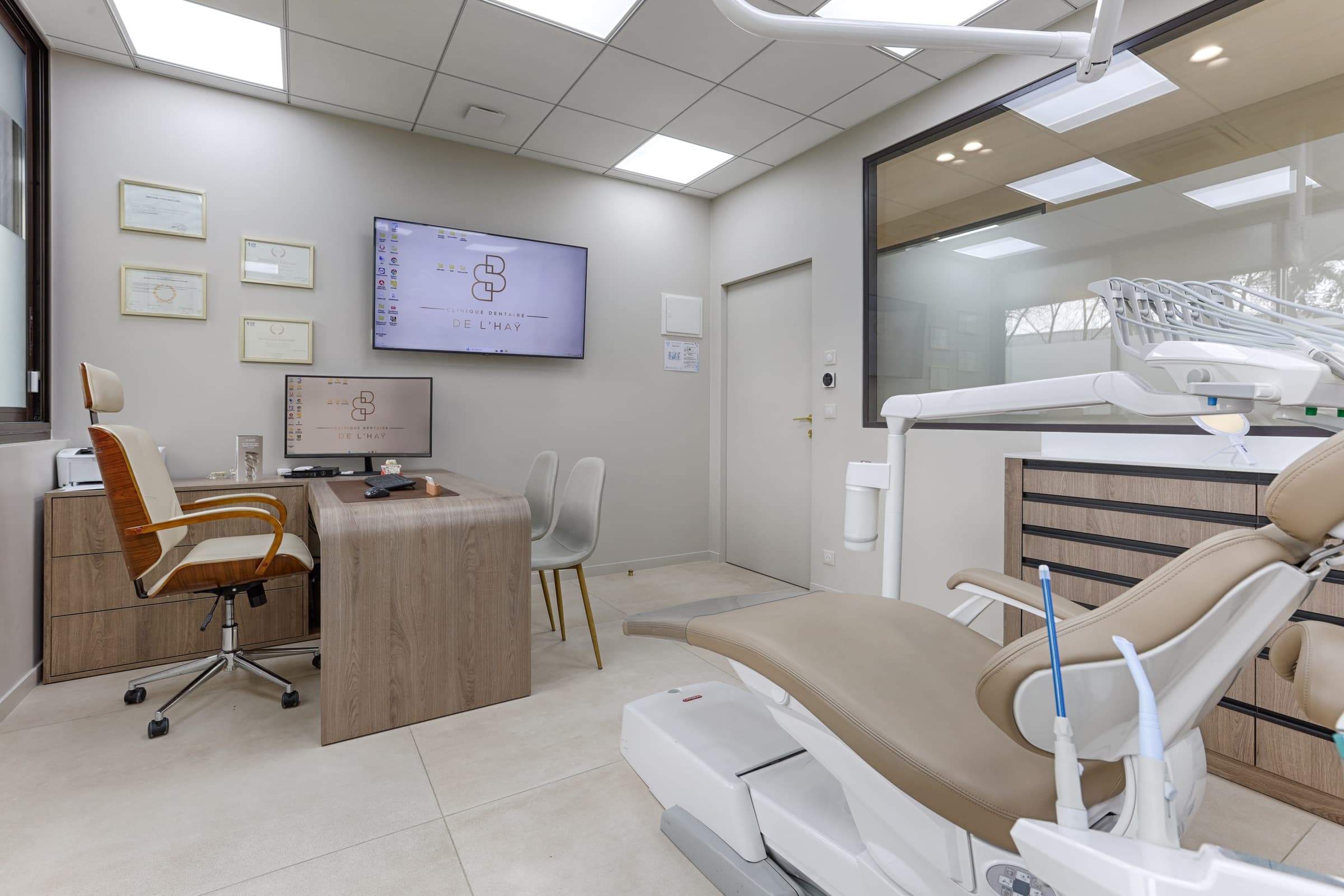Salle de soin équipée avec fauteuil dentaire beige, écran logo, bureau et certifications encadrées